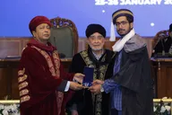 Receiving the university gold medal on the convocation stage, in graduation gown, from two university officials in ceremonial robes.